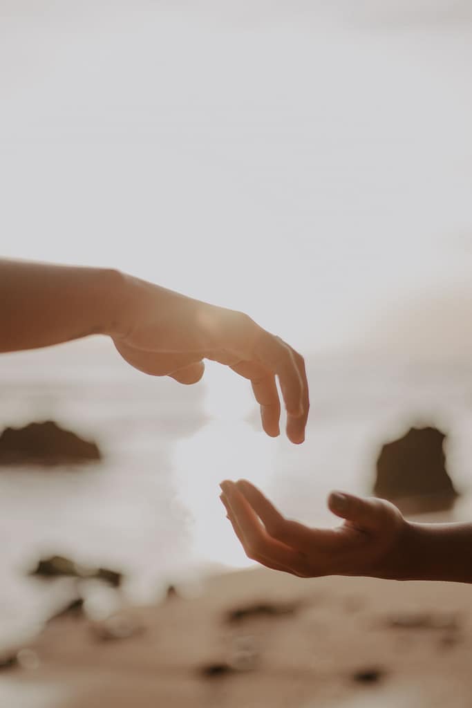 Close-up of two hands reaching towards each other with a serene beach background and soft lighting.