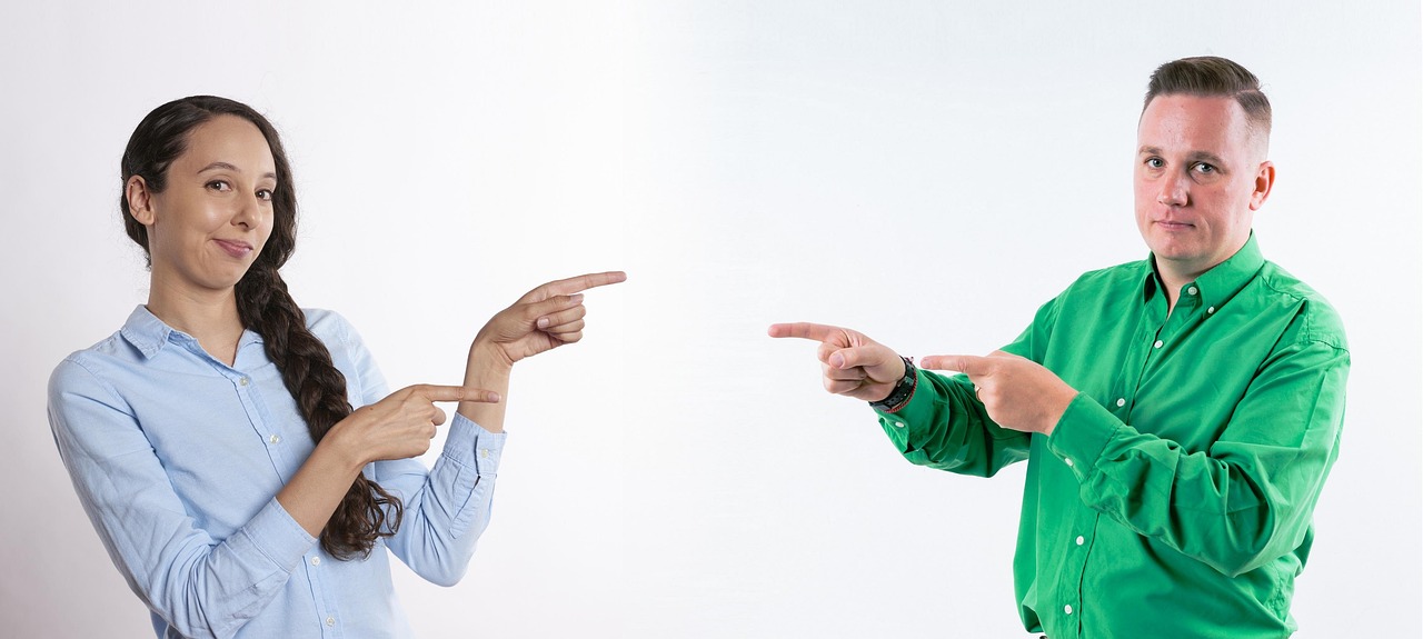 Two individuals, a woman in a light blue shirt and a man in a green shirt, are pointing in opposite directions against a plain white background.