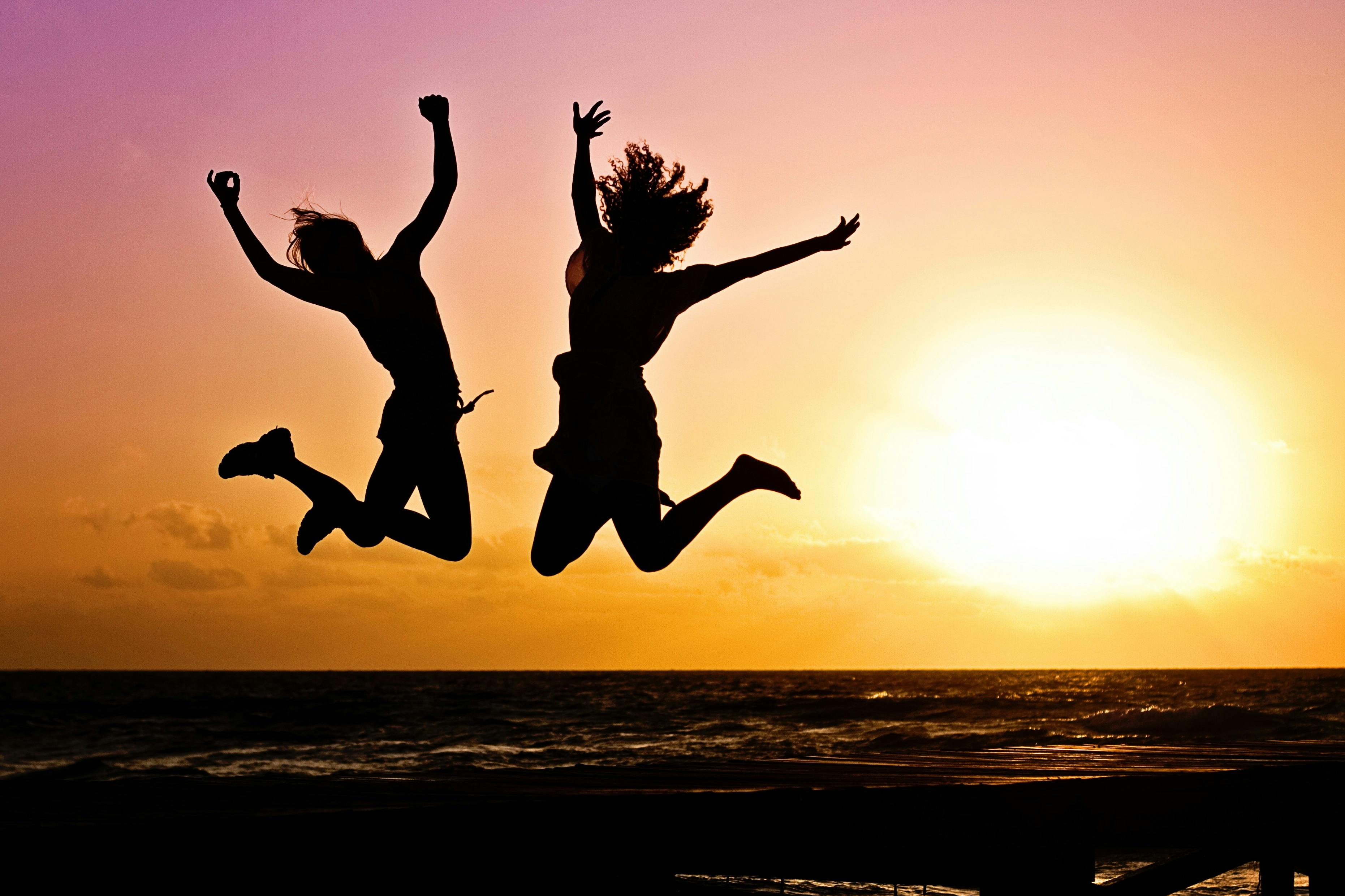 Two silhouetted figures jumping in joy against a vibrant sunset over the ocean.